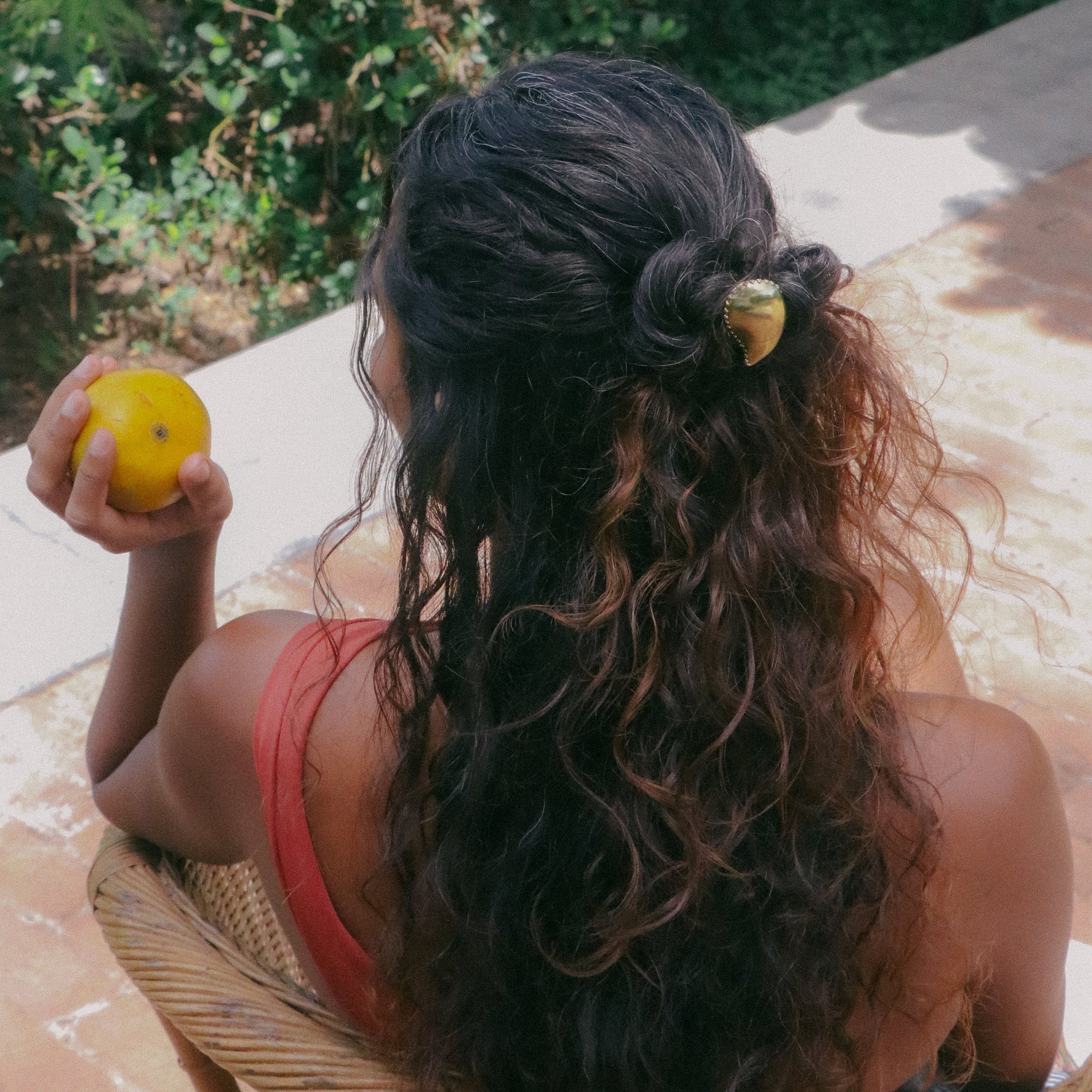 A woman with long, dark, curly hair is seen from behind, sitting in a wicker chair and holding a yellow fruit. Her hair is styled with a small gold hair accessory, and she is wearing a coral-red tank top.