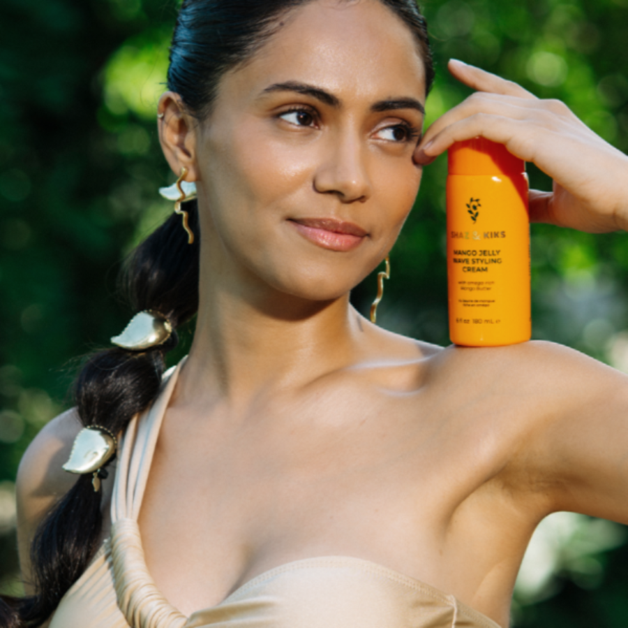 A woman outdoors holds a bright orange bottle of Richey's Mango Jelly Anti Dandruff Cream against her shoulder, with a blurred green natural background.